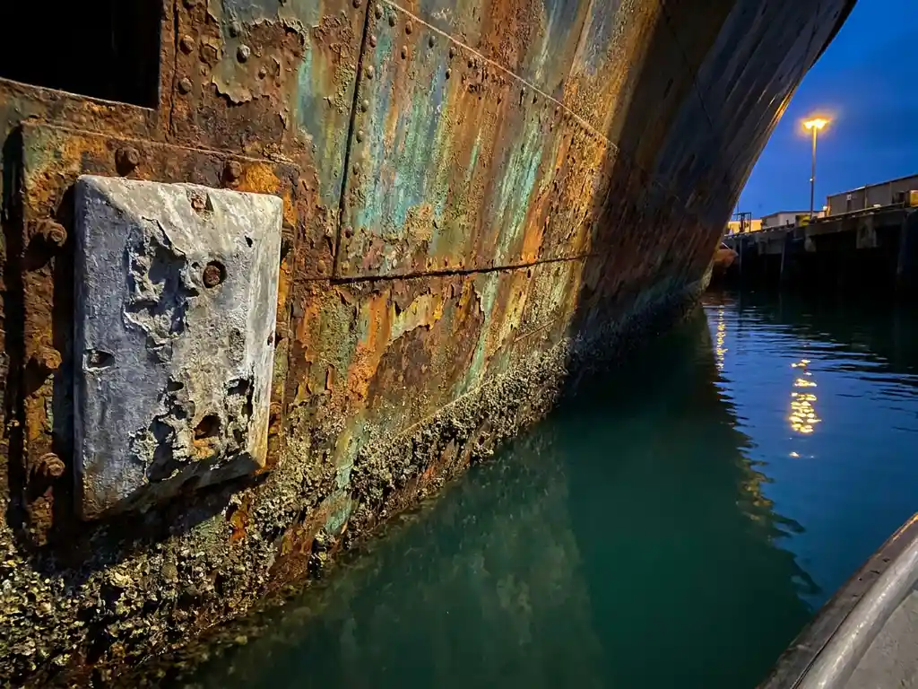 Verroest scheepsromp met groenblauw patina en uitgeputte zinken offerende anode boven donker havenwater, dramatisch zijlicht.