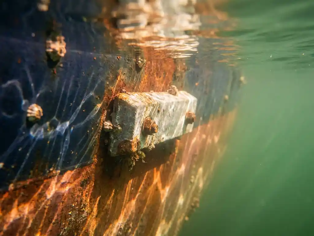 Zinkofferanode bevestigd aan verweerde stalen scheepsromp onder de waterlijn, omgeven door troebel groen havenwat